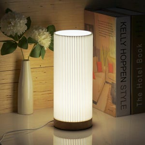 May include: A white cylindrical table lamp with a wood base and a white ribbed shade. The lamp is turned on and illuminating the room. The lamp is sitting on a table next to a stack of books.
