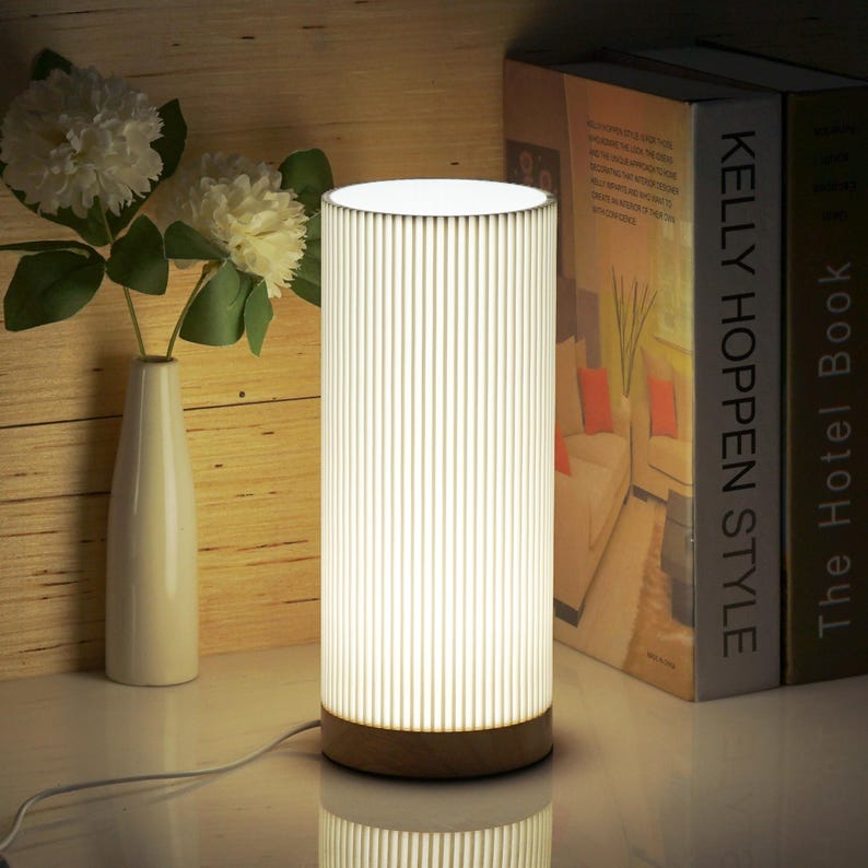 May include: A white cylindrical table lamp with a wood base and a white ribbed shade. The lamp is turned on and illuminating the room. The lamp is sitting on a table next to a stack of books.