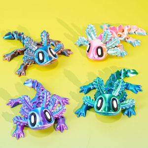 Puede incluir: Cuatro figuras de ajolote coloridas en azul, rosa, morado y verde con ojos blancos y negros se muestran sobre un fondo amarillo. Las figuras tienen una apariencia texturizada, impresa en 3D, con branquias y extremidades detalladas. Los ajolotes están en posición de gateo.