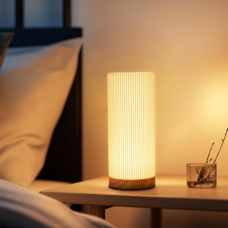 May include: A white bedside lamp with a wood base and a cylindrical shade with vertical lines. The lamp is turned on and casting a warm glow.