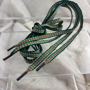 May include: A pair of multi-colored shoelaces. The laces are green, yellow, and red with black edges. The aglets are dark gray. The laces are knotted and arranged on a clear surface.