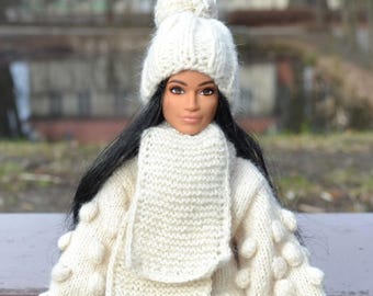 Puede incluir: Una muñeca con cabello largo y negro que lleva un gorro de punto color crema con un pompón, una bufanda de punto a juego y un suéter de punto color crema con detalles texturizados de pompones en las mangas. La muñeca tiene piel morena y ojos oscuros.
