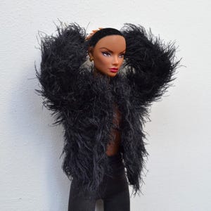 May include: A doll with a dark complexion and short dark hair wearing a black long-sleeved jacket made of a shaggy, textured material. The doll has gold-colored stud earrings, defined eyebrows, and pink lipstick, and is wearing black pants.