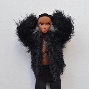 May include: A fashion doll in a black feathery jacket and black trousers. The doll has dark skin, dark hair, and gold earrings. The doll's arms are raised, and the jacket is open, showing the doll's chest. The background is plain white.