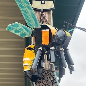 May include: A large, handcrafted metal bee sculpture with yellow and black stripes, blue-green wings, and black legs. The bee is mounted on a wooden post with a solar light and a bee house below.