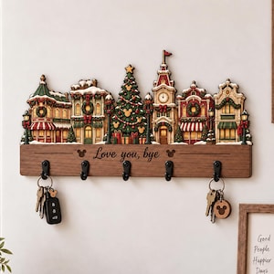 以下が含まれることがあります： 中央のクリスマスツリー、時計塔、店舗が描かれた、多色で層状のクリスマスビレッジシーンが特徴の木製壁掛けキーホルダー。5つの黒い金属製フックと、Love you, byeと刻印されたテキストが含まれています。