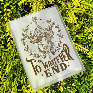 Op de afbeelding: Een doorzichtige, rechthoekige hoes met de woorden "To Whatever End" in een decoratief lettertype. De hoes heeft een bruine illustratie van een figuur met geweien, omgeven door een krans. De hoes staat tegen een achtergrond van groen gebladerte.