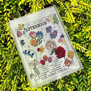 Puede incluir: Un bloque acrílico transparente rectangular con un diseño floral y el texto "All in the golden Afternoon". El diseño presenta ilustraciones coloridas de varias flores, incluyendo pensamientos, rosas y narcisos. El bloque está sobre un fondo de follaje verde.