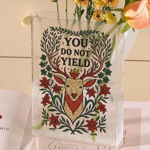 May include: A clear rectangular glass vase with the text "YOU DO NOT YIELD" and a deer illustration. The vase holds yellow and peach flowers. The deer illustration has red, green, and gold accents.