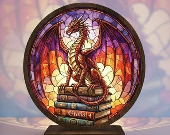 Op de afbeelding: Een gebrandschilderde lamp met een rode draak bovenop een stapel boeken. De lamp heeft een houten voet en een cirkelvormig ontwerp met levendige kleuren, waaronder rood, oranje en paars. De boeken dragen de naam "Olivia".