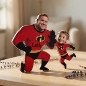 May include: Wooden cutouts of a man and a child dressed as superheroes, in red and black costumes. The man's cutout says "Daddy" and the child's says "Dash". They are on a wooden table with lavender sprigs.