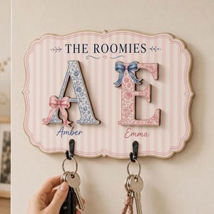 May include: A wall-mounted wooden key holder with a pink and white striped background. It features the text THE ROOMIES, large letters A and E with floral patterns and bows, and the names Amber and Emma. Two black metal hooks hold sets of keys.