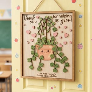 Könnte beinhalten: Ein rechteckiges Holzschild mit Aufhängeseil, das eine Topfpflanze mit grünem Laub und einem lächelnden Gesicht zeigt. Der Text lautet Thank you for helping us grow! sowie Love Miss Honey's Grade 1 Class 2026.