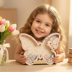 May include: A wooden butterfly-shaped keepsake box with the name "Isabella" on it. The box is light pink and filled with confetti and small decorative elements. A cartoon butterfly is on the right side of the box. A young girl is holding the box.