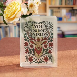May include: A clear rectangular vase with the words "YOU DO NOT YIELD" and a deer illustration. The deer is brown and red, surrounded by green leaves and red flowers. Yellow roses and white flowers are in the vase.