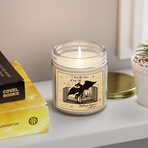 May include: A glass candle jar with a white candle inside. The jar has a label with a black and white illustration of a dragon flying out of an open book. The text on the label reads "I had the time of my life fighting dragons with you."