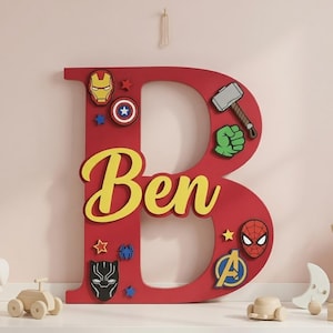 Puede incluir: Una gran letra "B" roja de madera decorada con emblemas de superhéroes y el nombre "Ben" en escritura amarilla. Los emblemas incluyen a Iron Man, Capitán América, el martillo de Thor y Spider-Man. Es un objeto decorativo.