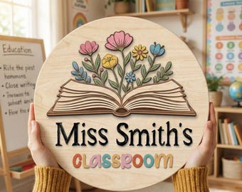 Pode incluir: Uma placa circular de madeira com um livro aberto cortado a laser de onde brotam flores coloridas. O texto "Miss Smith's" está gravado a preto e a palavra "classroom" aparece por baixo em letras 3D multicoloridas.