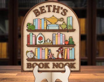 Może przedstawiać: Drewniany znak w kształcie półki na książki z napisem "BETH'S BOOK NOOK". Znak posiada trzy półki z kolorowymi ilustracjami książek i elementami dekoracyjnymi, takimi jak żaba, róża i imbryk. Stoi na białym drewnianym stojaku.