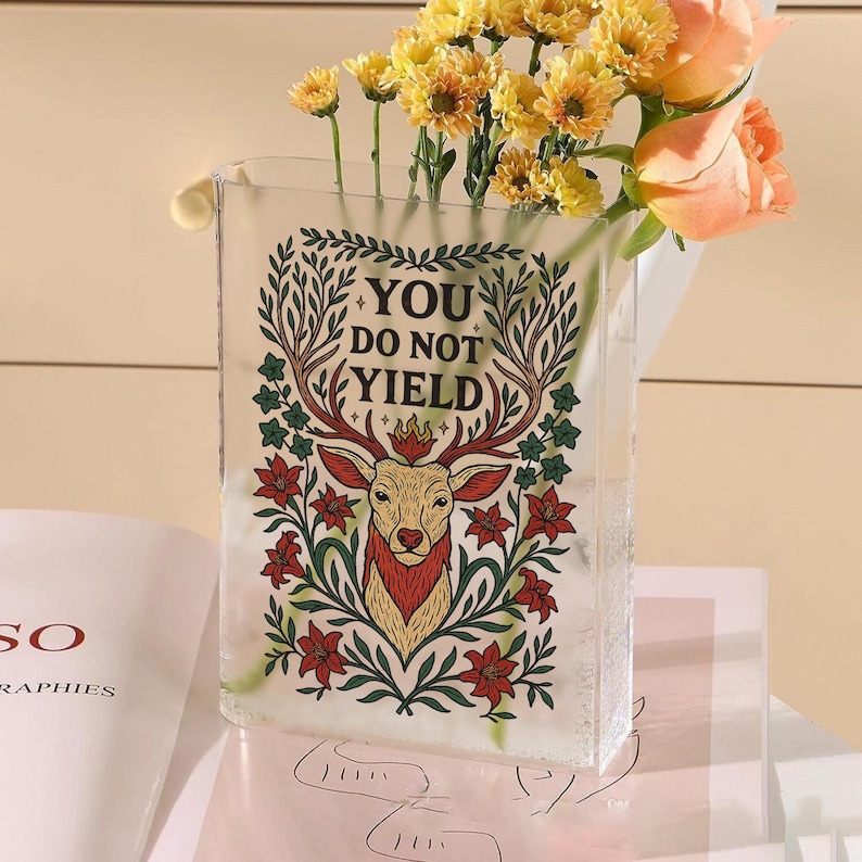 May include: A clear rectangular glass vase with the text "YOU DO NOT YIELD" and a deer illustration. The vase holds yellow and peach flowers. The deer illustration has red, green, and gold accents.
