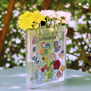 May include: A clear, rectangular vase with a floral design and the words "All in the golden AFTERNOON." The vase holds yellow and white flowers. The design includes illustrations of various flowers with their names.
