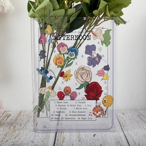 May include: Clear rectangular vase with a floral design and the text "Afternoon." The vase features illustrations of various flowers, including sweet peas, pansies, and roses. Green foliage is inside the vase.