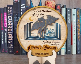 May include: A wooden round plaque with a black dragon flying out of an open book. The plaque has the text "I had the time of my life fighting dragons with you Olivia's Library" and a decorative floral design. The plaque is on a wooden stand.