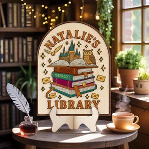 Puede incluir: Letrero de madera con el texto "NATALIE'S LIBRARY" e ilustraciones de libros, un búho y un castillo. El letrero está apoyado en un soporte de madera. Una pluma y un tintero están sobre una mesa a la izquierda y una taza de té a la derecha.
