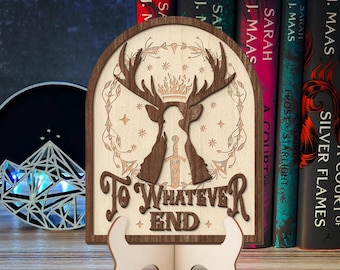 Op de afbeelding: Een houten boekenlegger met een silhouet van een hert met geweien en een kroon, met de woorden "To Whatever End" eronder gegraveerd. De boekenlegger staat op een houten tafel met een stapel boeken erachter.