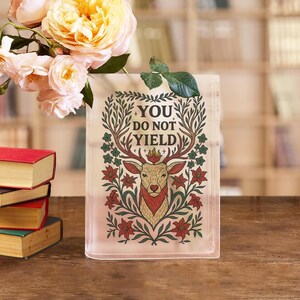 May include: A rectangular glass vase with the text "YOU DO NOT YIELD" and a deer illustration. The deer has antlers and is surrounded by floral designs in red, green, and gold. A bouquet of yellow and pink roses sits to the left of the vase.