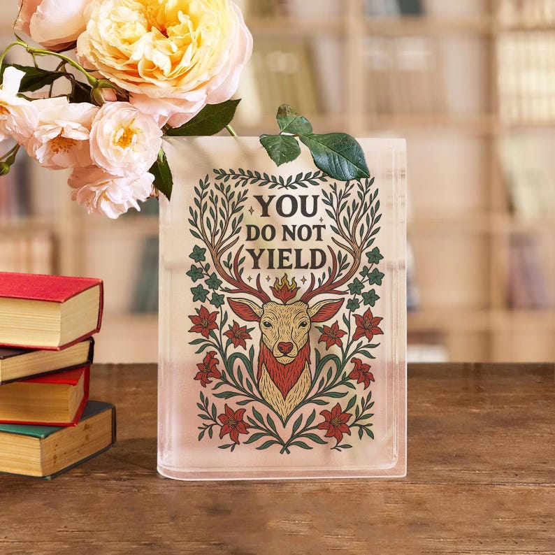 May include: A rectangular glass vase with the text "YOU DO NOT YIELD" and a deer illustration. The deer has antlers and is surrounded by floral designs in red, green, and gold. A bouquet of yellow and pink roses sits to the left of the vase.