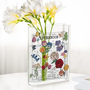 May include: A clear acrylic vase with a floral design and the words "All in the golden AFTERNOON." The vase holds white and yellow flowers. The vase has a decorative floral design with numbered flower names.