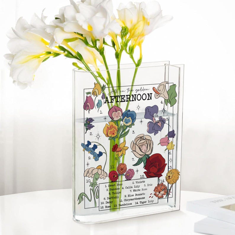 May include: A clear acrylic vase with a floral design and the words "All in the golden AFTERNOON." The vase holds white and yellow flowers. The vase has a decorative floral design with numbered flower names.