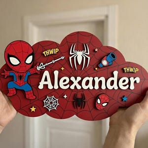 Könnte beinhalten: Ein rotes Holzschild in Wolkenform mit Spider-Man-Motiv. Das Schild enthält den Namen "Alexander" in weißen Buchstaben sowie verschiedene Spider-Man-Grafiken, wie ein Spinnennetz, eine Spinne und einen Netzwerfer.