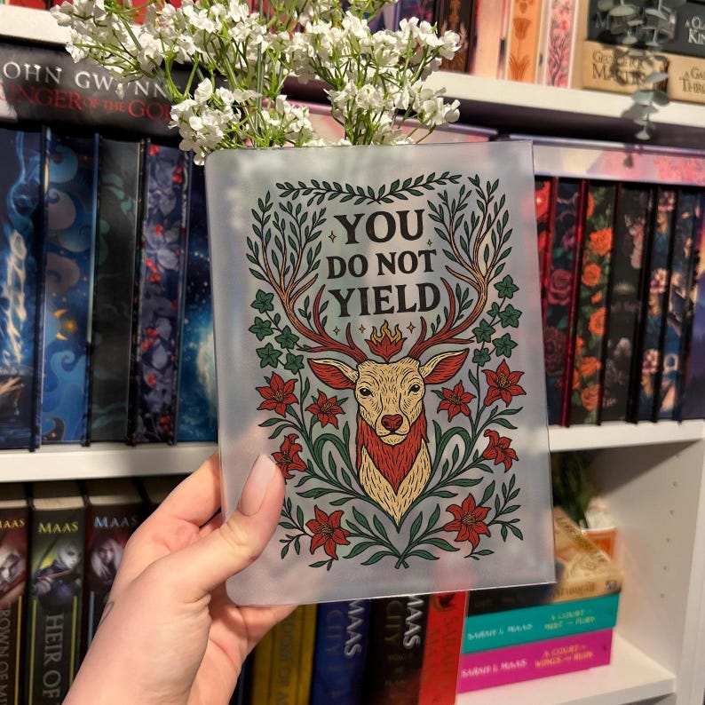 May include: A clear book cover with the text "YOU DO NOT YIELD" and a deer illustration. The deer has red antlers and is surrounded by red flowers and green leaves. The cover is held in front of a bookshelf.