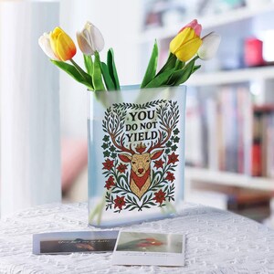 May include: A clear rectangular vase with a decorative deer design and the text "YOU DO NOT YIELD". The vase holds yellow, white, and pink tulips. Two small cards are on the table in front of the vase.