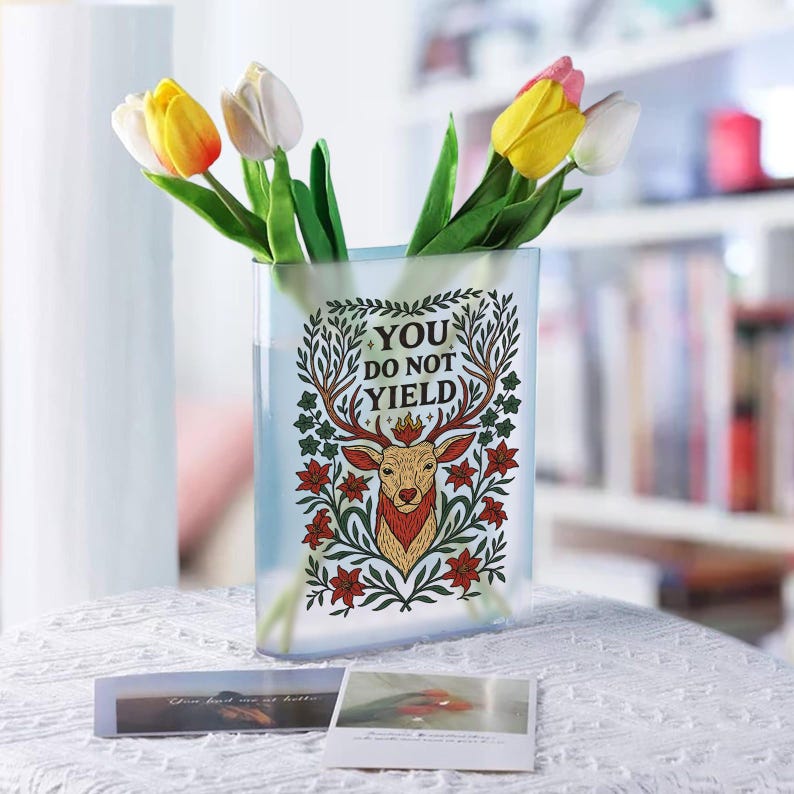 May include: A clear rectangular vase with a decorative deer design and the text "YOU DO NOT YIELD". The vase holds yellow, white, and pink tulips. Two small cards are on the table in front of the vase.