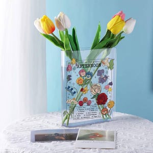May include: A clear rectangular vase with a floral design and the text "All in the golden Afternoon" holds colorful tulips. The vase features illustrations of various flowers and a list of flower names. The vase sits on a white surface.