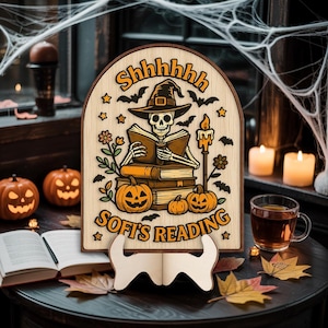 Peut inclure: Panneau en bois sur le thème d'Halloween avec un squelette lisant un livre, portant un chapeau de sorcière, entouré de citrouilles et de chauves-souris. Le panneau indique "Shhhhhhh Sofi's Reading". Le panneau est présenté sur un support en bois.