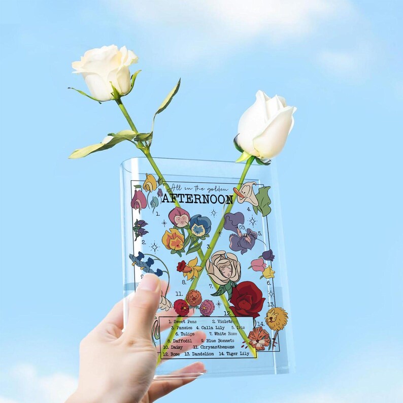 May include: A clear, rectangular vase with a floral design and the text "All in the golden AFTERNOON" holds two white roses. The vase features illustrations of various flowers and their names. The background is a bright blue sky.