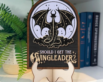 May include: A wooden sign with a black background and a white silhouette of a dragon with stars. The text on the sign reads "Should I get the Wingleader?"