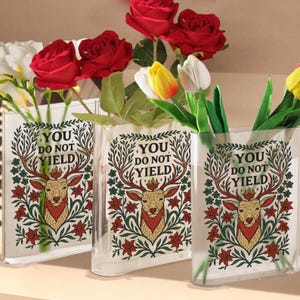 May include: Three clear glass vases with a deer and floral design, and the text "YOU DO NOT YIELD". The vases hold red roses, yellow and white tulips, and other greenery. The vases are rectangular and have a slight curve.