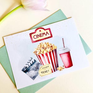 Könnte beinhalten: Eine weiße Karte mit einem rot-weiß gestreiften Popcorn-Eimer, einem roten Becher mit Strohhalm und einer Filmklappe. Die Karte hat auch ein gelbes Ticket mit dem Wort "TICKET" darauf und ein rotes Schild mit weißen Sternen und dem Wort "CINEMA" darauf.