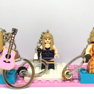 Puede incluir: Tres figuras estilo Lego de una cantante, cada una con un atuendo y un micrófono diferentes. Una figura sostiene una guitarra rosa. Las figuras están unidas a llaveros y se asientan sobre una base rosa y azul.