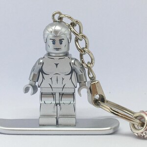 Könnte beinhalten: Eine silberfarbene Lego-Figur eines Superhelden mit detailliertem Torso und Kopf, die auf einer silbernen Plattform steht. Angebracht sind eine silberne Kette und ein rosa-silberner Anhänger. Die Figur ist ein Schlüsselanhänger.
