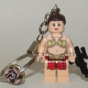 Puede incluir: Un llavero Lego de la princesa Leia de Star Wars. La figura lleva un top de bikini dorado y una falda roja. Lleva un bláster en su mano derecha. El llavero está unido a un anillo plateado.