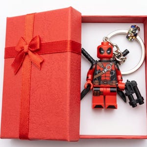 Può includere: Una scatola regalo rossa con fiocco in nastro abbinato contenente un portachiavi del personaggio Deadpool. Il portachiavi presenta una figura in plastica rossa e nera con due spade nere sulla schiena e un'arma in plastica nera in mano.