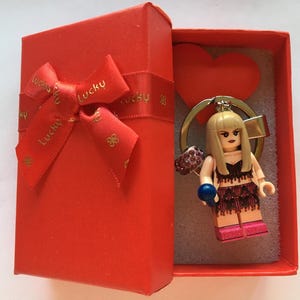 Puede incluir: Una caja de regalo roja con lazo y cinta a juego, que contiene un llavero con figura estilo Lego. La figura tiene cabello rubio, un top oscuro y una falda rosa. Una forma de corazón rojo es visible en el fondo.