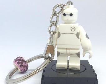 May include: A white and gray Baymax-inspired keychain featuring a Lego-style figure with black eyes and a circular chest emblem. The keychain includes a silver chain, a pink and purple beaded charm, and a silver key ring.