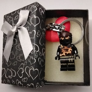 May include: A black gift box with a silver bow, containing a Lego-style ninja keychain. The ninja figure is black with tan and brown accents. A red heart-shaped charm and a silver keyring are also included.